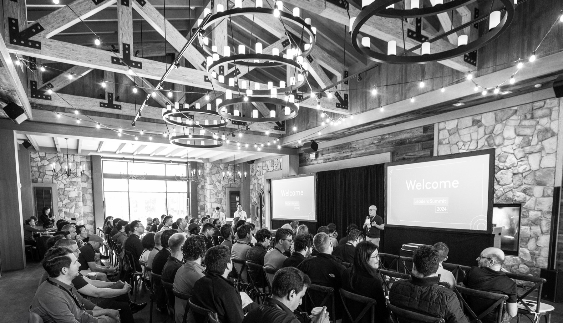 A leaders summit underway in a timber-beamed lodge, chandeliers and string lights above a seated audience